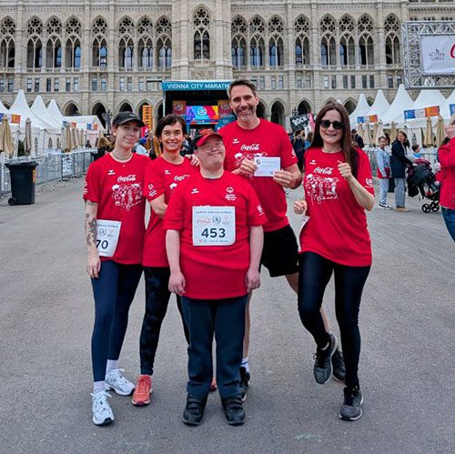 Künstler David Cheng in der Mitte, rechts und links von ihm stehen VOI fesch Mitarbeiter*innen, alle fünf Personen tragen das Coca-Cola Shirt für den Inclusion Run mit dem VOI fesch Design des Künstlers David Cheng, im Hintergrund sind Menschen am Rathausplatz zu sehen