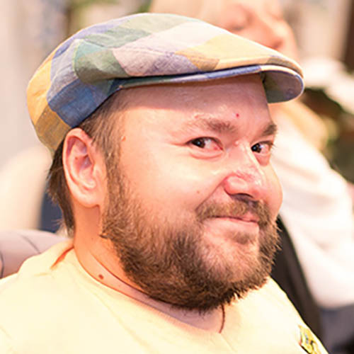 Artur Belja, VOI fesch Künstler; er schaut seitlich in die Kamera und lächelt, er trägt eine Kappe und einen Vollbart