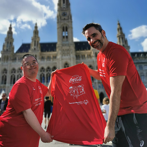Coca-Cola Shirt für den Inclusion Run mit VOI fesch Design von Künstler David Cheng, im Bild ist links der Künstler und rechts Helmuth von VOI fesch, beide halten ein Laufshirt in den Händen, damit das Design sichtbar wird; im Hintergrund ist das Rathaus in Wien zu sehen