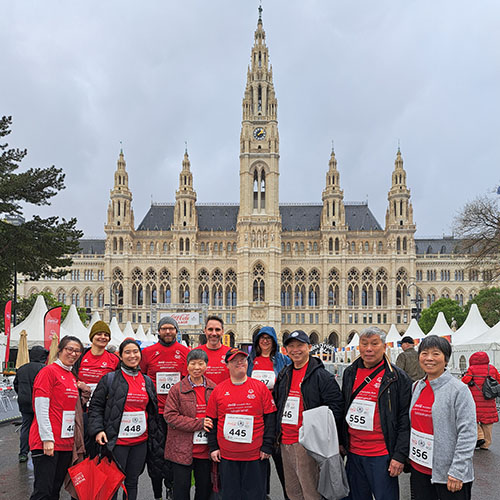 Gruppenbild mit Menschen, unter anderem dem Künstler David Cheng in der Mitte, alle tragen das rote Laufshirt mit seinem Design, entworfen für den Coca Cola Inclusion Run; im Hintergrund ist das Rathaus in Wien zu sehen