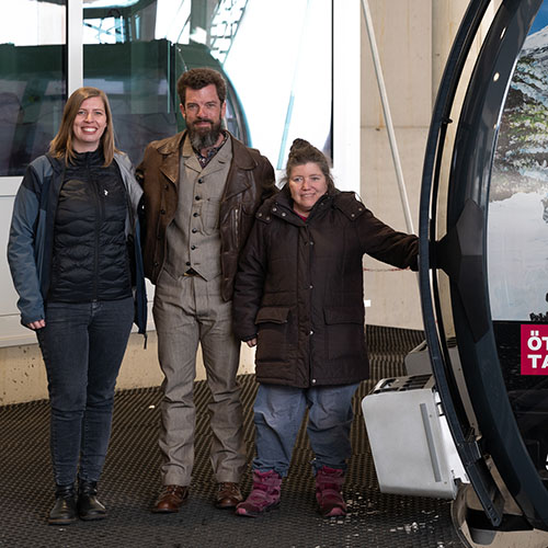 Foto in der Talstation der Rosskarbahn, zwei Frauen und ein Mann stehen neben einer Gondel, von der nur die Tür und ein kleiner Teil zu sehen sind; alle drei Personen lächeln in die Kamera