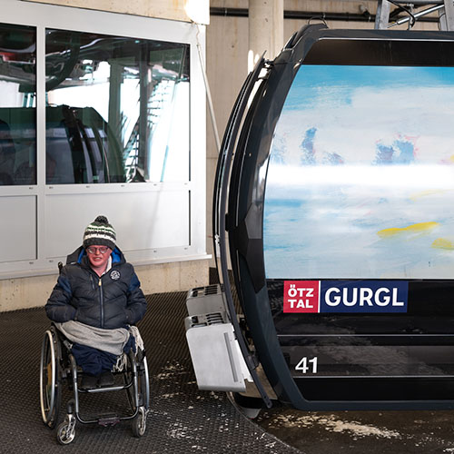 im Bild Künstler Florian Haider neben der Gondel, die mit seinem Kunstwerk beklebt ist, der Künstler trägt eine Haube, Brillen und eine warme Jacke, er sitzt im Rollstuhl