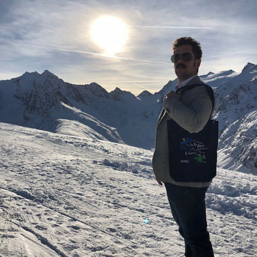 Mann steht seitlich zur Kamera im Vordergrund, Tasche hängt an seiner Schulter, er trägt eine Sonnebrille und einen Bart, blickt in die Kamera; im Hintergrund ist schneebedeckter Boden, Berge mit Schnee und die Sonne zu sehen