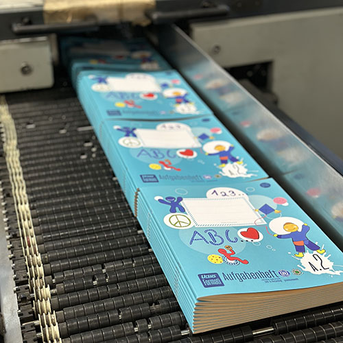 Formatwerk Heft in Produktion: ertige Aufgabenhefte im VOI fesch Design liegen gestapelt hintereinander, aufgelegt auf einer Maschine mit Rollen – in der Druckerei.