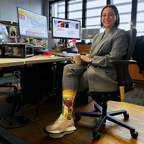 Frau sitzt an einem Schreibtisch im Bürosessel. Sie ist zur Kamera gedreht und lächelt, dass ihre Zähle sichtbar sind. Sie trägt eine graue Hose und einen grauen Blazer. In der Hand hält sie eine Kaffee-Tasse. Die Hosenbeine sind weit hochgezogen, somit werden die bunten Socken im VOI fesch Design sichtbar. Sie trägt helle Turnschuhe, im Hintergrund sind Computer-Monitore und eine Fensterfront.