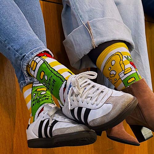 Zwei Menschen sitzen erhöht nebeneinander. Nur ihre Beine und Füße sind zu sehen. Sie tragen die selben bunten Socken im VOI fesch Design von Künstler Albert Masser.
