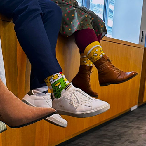 Zwei Menschen sitzen auf einer Fensterbank nebeneinander. Nur ihre Beine und Füße sind zu sehen. Sie tragen alle die selben bunten Socken im VOI fesch Design von Albert Masser. Aus dem vorherigen Foto sind noch die Schuhe einer weiteren Person zu sehen.
