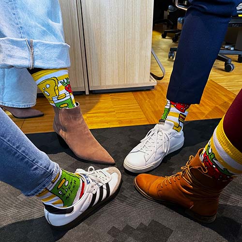 Menschen haben ihre Füße mit der Spitze, den Zehen voran, aneinander gestellt. Es sind nur die Beine und Füße sichtbar. Alle haben die Hosen hochgezogen damit die bunten Socken im VOI fesch Design zu sehen sind. Sie stehen alle auf einem grauen Teppich.