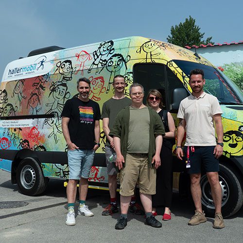 Eine Gruppe von fünf Menschen steht vor einem bunten Fahrzeug mit VOI fesch Design. Alle schauen und lächeln in die Kamera, alle haben Sommerkleidung an, kurze oder knöchellange Hosen. Im Hintergrund ist ei Stück einer Plakatwand sichtbar.