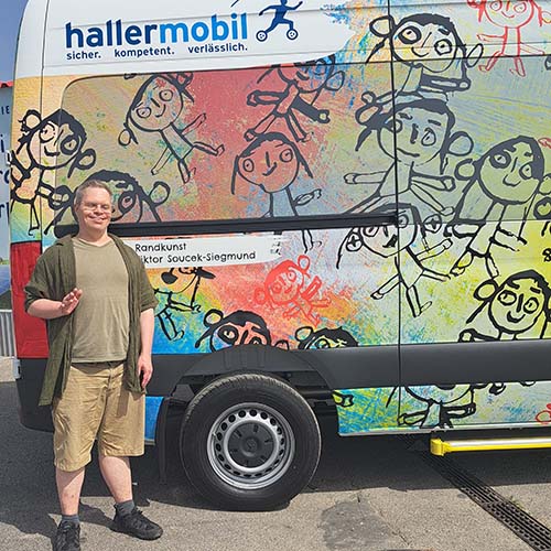 Ein Mann steht vor einem bunten Fahrzeug im VOI fesch Design. Er lacht und winkt in die Kamera. Er trägt eine Brille, kurze Haare und hat ein T-Shirt, ein Hemd darüber und eine kurze Hose an.